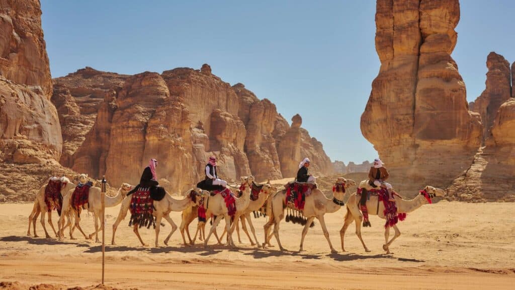 A small camel train with riders in traditional dress crossing the sandstone valley of AlUla, Saudi Arabia, on the old caravan route.