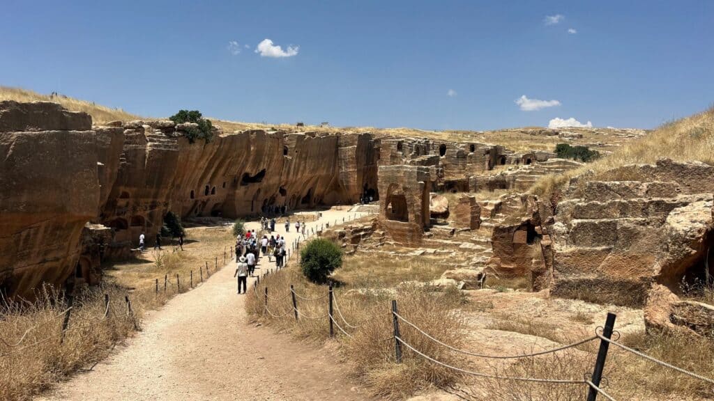 Wide view of the rock-cut quarry-necropolis at Dara, southeastern Turkey, with visitors walking a path below cliff faces containing tomb chambers.