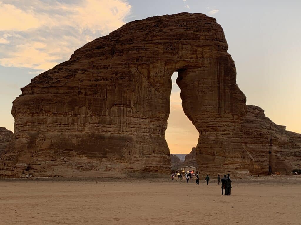Elephant Rock (Jabal AlFil) at sunset in AlUla, Saudi Arabia, its trunk-shaped arch towering over visitors on the sand.