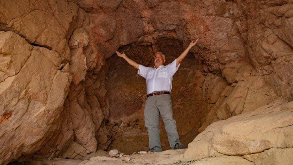 A man stands upright, arms raised joyfully, inside the Cave of Elijah on Jabal Maqla, the chamber rising well above his head.