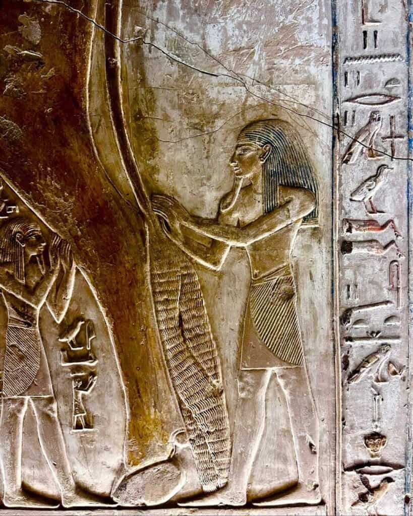 Painted relief in the tomb of Seti I showing two figures in long wigs and pleated kilts with arms raised, hands against the underside of a large bull, beside a painted column of hieroglyphs