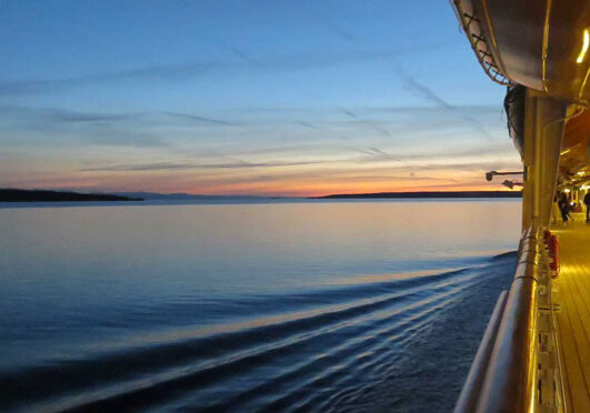 A long view of a cruise ship deck at sunset with shore in the distance.